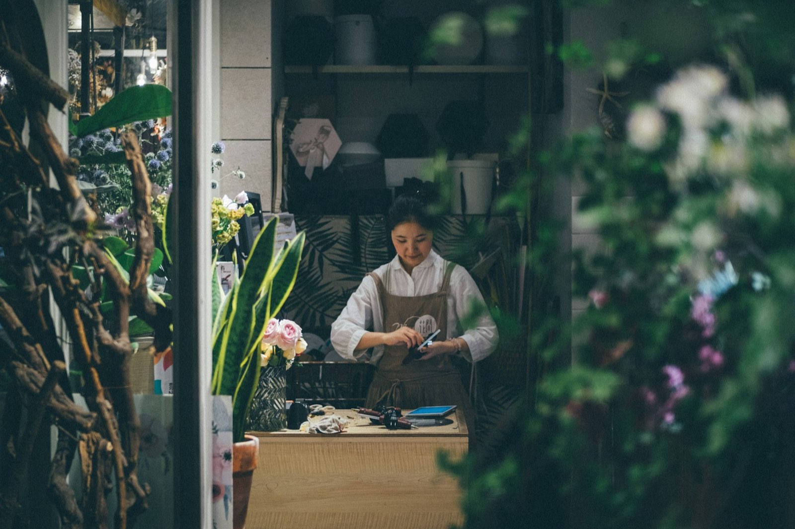 A woman working at the counter of a plant and goods shop, framed by green plants and flowers.